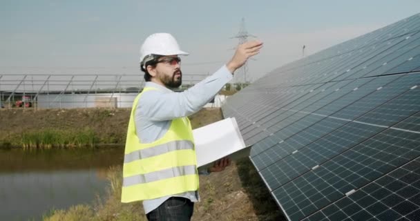 Positive adult man in uniform checking photovoltaic panels and making notes in journal during work on solar power station. checking the panels at solar energy installation. - Video