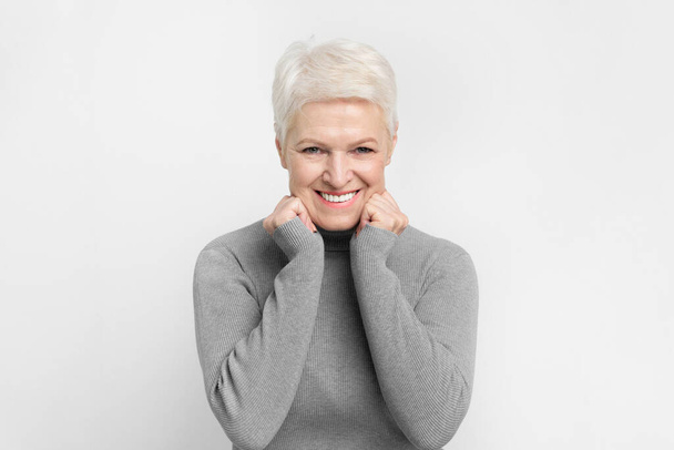 Radostné starší Evropanka s hravým gestem třít si tváře na šedém pozadí, ideální pro s3niorlife štěstí - Fotografie, Obrázek