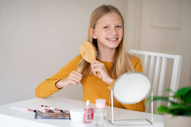 Menina adolescente alegre escovando seu cabelo longo, sentado a uma mesa com um espelho e cosméticos, desfrutando de cuidados de beleza - Foto, Imagem