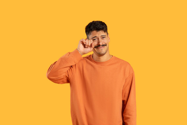 Slzící emocionální muž s knírkem v oranžovém svetru otírá slzu na žlutém pozadí - Fotografie, Obrázek