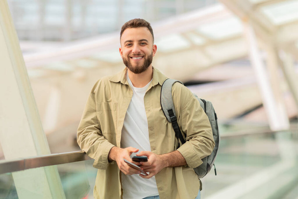Havalimanında beklerken elinde pasaport ve akıllı telefon tutan neşeli genç bir adam. Mutlu milenyum erkekleri yalnız seyahat ediyor. Yaklaşan yolculuk için heyecanlı. - Fotoğraf, Görsel