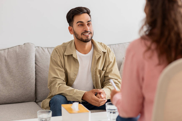 Image shows a young man with a beard patient engaging in a conversation with a woman therapist seated on a sofa - Photo, Image