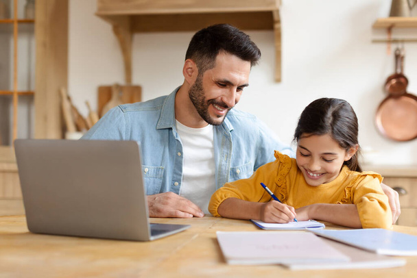 Улыбающийся мужчина ведет ребенка своим исследованием в теплой домашней обстановке, с ноутбуком и книгами на столе - Фото, изображение