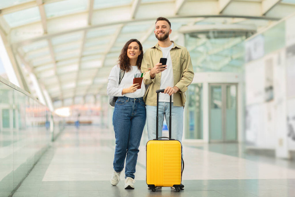 Mężczyzna i kobieta przechadzają się po ruchliwym terminalu lotniskowym, niosąc bagaż i szukając bramy startowej.. - Zdjęcie, obraz