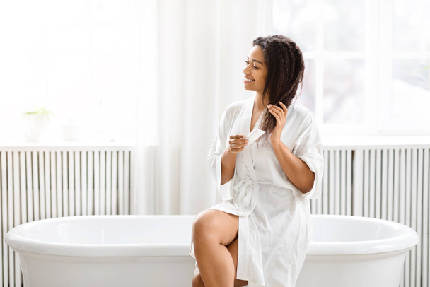 Tyylikkäässä ja minimalistisessa ympäristössä afroamerikkalainen nainen suorittaa henkilökohtaisen grooming-rutiinin kylpyammeen lähellä. - Valokuva, kuva