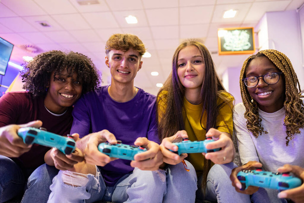 Μια χαρούμενη ομάδα εφήβων μοιράζεται μια διασκεδαστική στιγμή παίζοντας βιντεοπαιχνίδια σε ένα τοπικό κέντρο τυχερών παιχνιδιών. - Φωτογραφία, εικόνα