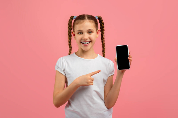 A happy girl points at her smartphone screen, looking at the camera with a smile on a pink background - Photo, Image