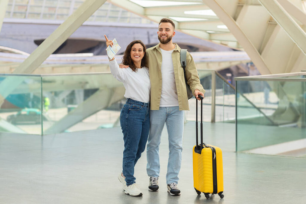 Fröhliches Paar, das mit Gepäck im Flughafenterminal steht und wegzeigt, lächelnder Mann und Frau mit gelbem Koffer, die am Gate auf ihren Flug warten und gemeinsam die Reise genießen - Foto, Bild