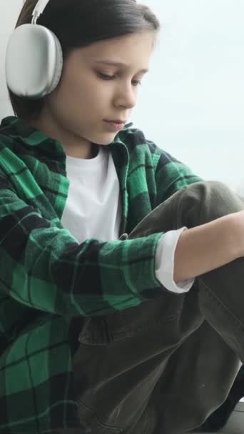Muotokuva onneton ja suru valkoihoinen nuori koulupoika kuuntelee musiikkia kuulokkeet ikkunalaudalla kotona. Henkisen stressin käsite. Pystysuora video. - Materiaali, video