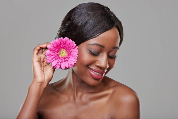 Γυναίκα, περιποίηση της επιδερμίδας και χαρούμενη με λουλούδι στο στούντιο για ομορφιά, βιολογικά καλλυντικά και μακιγιάζ από γκρι φόντο. Κορίτσι, μοντέλο και αφρικανικό πρόσωπο με ροζ μαργαρίτα, σκέψη και οικολογία με φυσική λάμψη. - Φωτογραφία, εικόνα