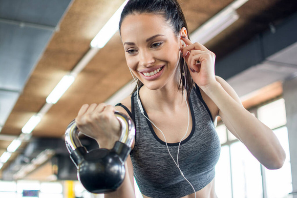 Low angle view of smiling sporty girl with earphones lifting kettlebell in the gym - Photo, Image