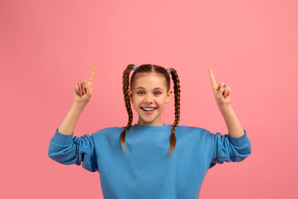 Joyeux jeune fille portant un pull bleu avec des cheveux tressés pointant joyeusement vers le haut sur un fond rose - Photo, image