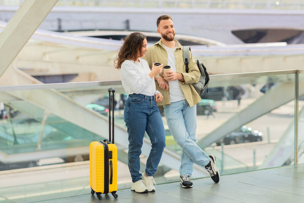 Hombre y mujer con café para llevar esperando vuelo en el aeropuerto, sonriente pareja joven con equipaje de pie en la terminal, charlando y riendo, emocionado por el próximo viaje - Foto, imagen