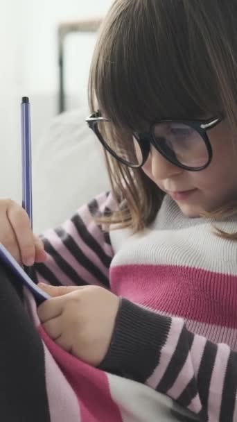 Retrato de uma menina caucasiana pré-escolar focada desenho ou escrita em caderno no sofá em casa. Conceito de aprendizagem ou imaginação. Vídeo vertical. - Filmagem, Vídeo