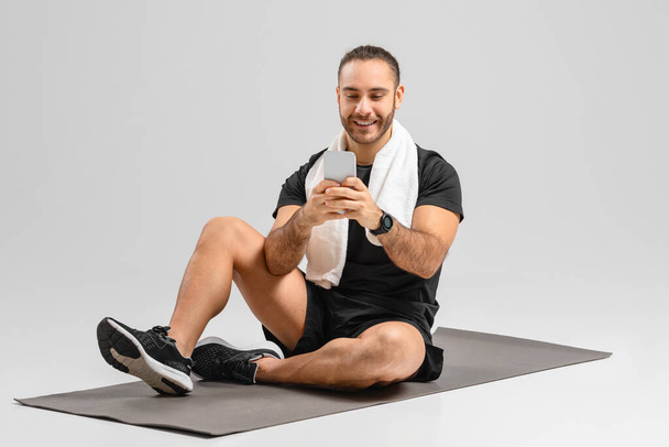 Χαρούμενος άνθρωπος χρησιμοποιώντας ένα smartphone, ενώ κάθεται σε ένα στρώμα γυμναστικής σε ένα περιβάλλον στούντιο άσκησης, ωραία εφαρμογή - Φωτογραφία, εικόνα