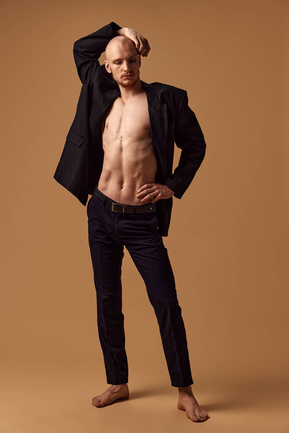 Shirtless young athletic man with elegant trousers and jacket showing casual sophistication against sandy studio background. Concept of mens health, self care, fashion and beauty, healthy lifestyle. - Foto, imagen