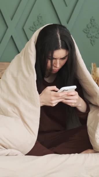 Rozrušená běloška zabalená do deky telefonem v posteli v domácí ložnici. Psaní nebo čtení on-line diskusní koncept. Svislé video. - Záběry, video