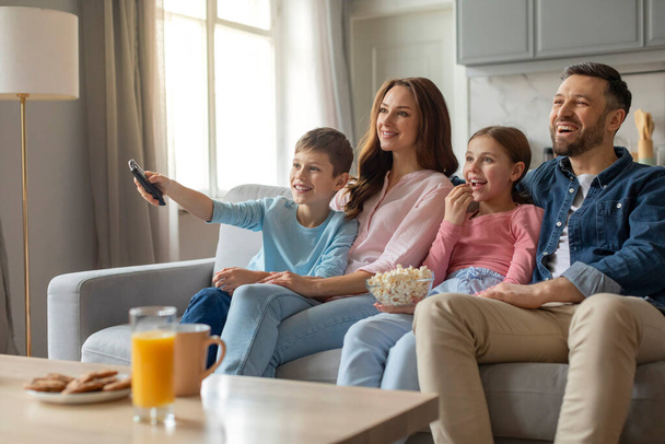 Веселая семья с двумя детьми, сидящими на диване и нажатием пульта дистанционного управления во время просмотра телевизора дома - Фото, изображение