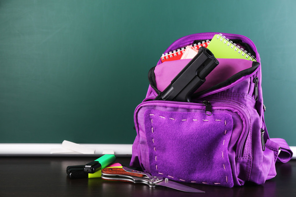 Gun in school backpack on wooden desk, on blackboard background - 写真・画像