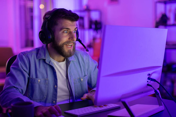 Homme avec écouteurs intensément concentré sur un écran d'ordinateur, entouré d'une pièce maussade éclairée bleue à la maison - Photo, image