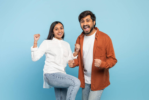 An exuberant Indian couple with fists raised celebrating victory or success together with joy on blue - Photo, Image