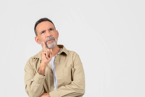 Nachdenklicher älterer Mann in lässiger Kleidung blickt mit dem Finger auf die Wange und zeigt tief in Gedanken oder Entscheidungen, isoliert auf weißem Hintergrund - Foto, Bild