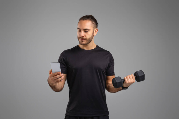 Egy férfi felemel egy súlyzót, miközben egyidejűleg ellenőrzi az okostelefonját, illusztrálva a fitness mobilalkalmazást - Fotó, kép