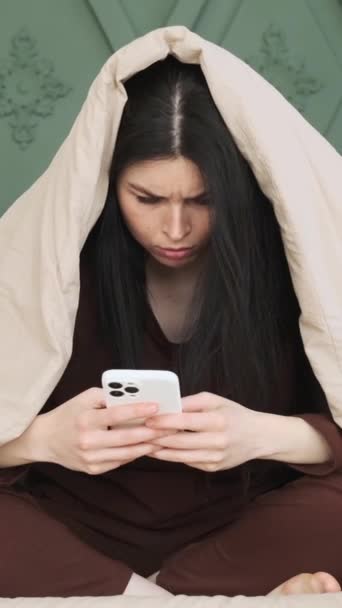 Femme caucasienne bouleversée ou ennuyée enveloppée dans une couverture, messagerie en ligne dans les médias sociaux en utilisant le téléphone sur le lit et tomber. Concept de temps libre ennuyé. Vidéo verticale. - Séquence, vidéo
