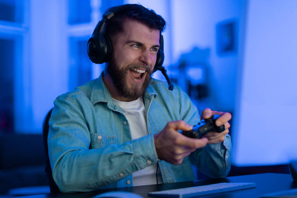 Hombre barbudo feliz con un controlador de juego siente emoción durante un juego, luz azul llenando la habitación - Foto, imagen