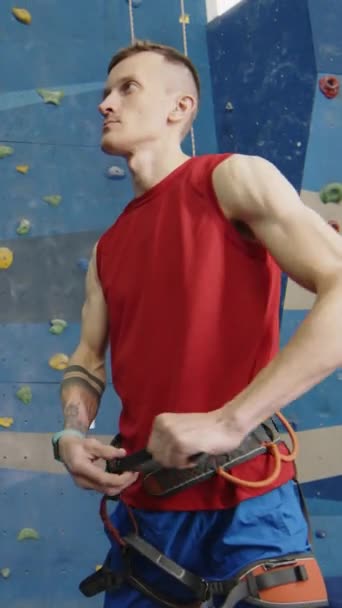 Tiro vertical de hombre musculoso con prótesis de miembro inferior de pie en el gimnasio interior y sujeción de equipo de escalada alrededor de la cintura - Imágenes, Vídeo