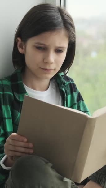 Gericht Kaukasische jongen zittend op vensterbank en boek lezen thuis. Ontspannen met literatuur en Home library concept. Verticale video. - Video