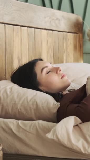Boldog és mosolygó kaukázusi nő ébred fel reggel, nyújtózkodik és fekszik az ágyban otthon, családi kikapcsolódás. Függőleges videó. - Felvétel, videó