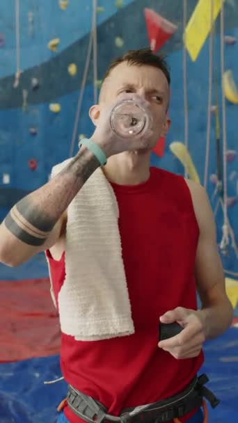 Plan vertical de l'homme fatigué en tenue de sport debout dans la salle de sport d'escalade, l'eau potable et essuyant la sueur du visage avec une serviette pendant la pause - Séquence, vidéo