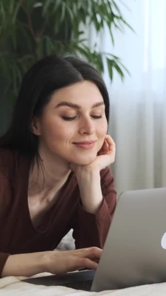Happy Caucasian woman using laptop while lying in bed, browsing internet or messaging online in social media. Remote communication concept. Vertical video. - Footage, Video