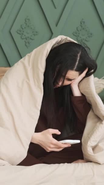 Donna caucasica preoccupata che scrive nell'app di social media online, usando il telefono seduto sul letto e avvolto in una coperta. Spendere passatempo concetto. Video verticale. - Filmati, video
