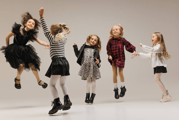 Leuke, coole houding kleine meisjes, elementaire studenten springen van vreugde en geluk tegen grijze studio achtergrond. Concept van de kindertijd, kinderen mode en schoonheid, stijl, school winkelen. Zwarte vrijdag - Foto, afbeelding