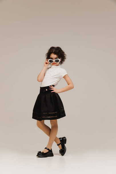 Γοητευτικό κορίτσι σε σικ μαύρη φούστα και λευκό τοπ, ποζάροντας παιχνιδιάρικα με τεράστια γυαλιά ηλίου σε γκρι φόντο στούντιο. Έννοια της παιδικής ηλικίας, τη μόδα και την ομορφιά των παιδιών, το στυλ, τα ψώνια του σχολείου. - Φωτογραφία, εικόνα