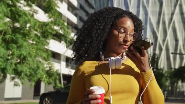 Ευτυχισμένη νεαρή μαύρη γυναίκα με γυαλιά και ακουστικά, έχοντας μια hands-free συνομιλία στο κινητό τηλέφωνο με καφέ στο χέρι. - Πλάνα, βίντεο