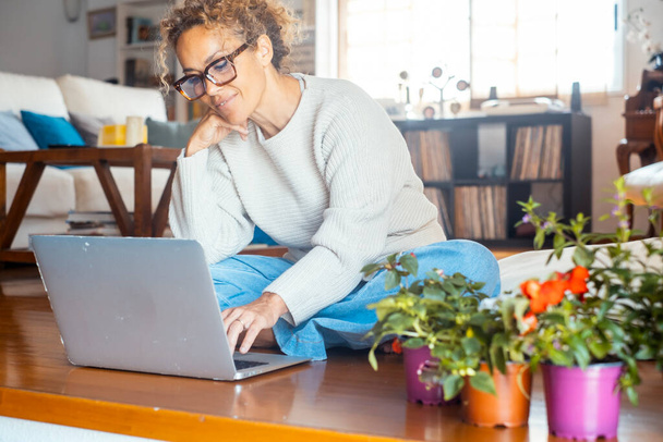 Eine erwachsene Frau, die allein zu Hause am Laptop arbeitet, sitzt auf dem Holzboden. Drahtlose Verbindung und Technologie. Weibliche moderne Menschen nutzen Computer, um das Internet zu durchsuchen und online einzukaufen - Foto, Bild