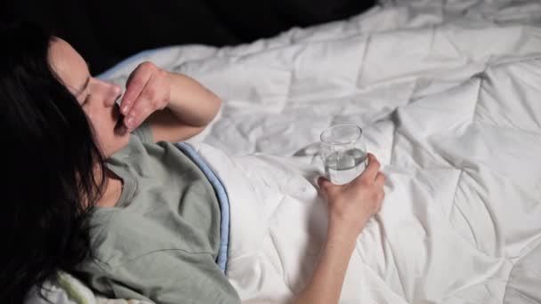 молода брюнетка спокійно лежить на ліжку, приймаючи її призначені ліки зі склянкою води її ніжний акт ковтання таблеток, рутина, яку вона старанно слідує за її благополуччям - Кадри, відео