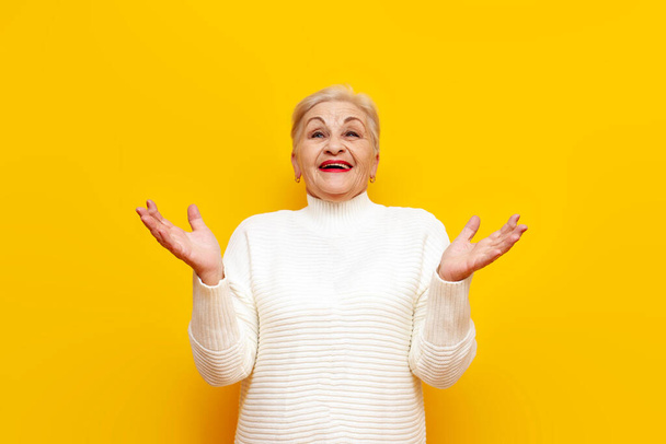 iloinen yllättynyt vanha mummo valkoisessa puserossa nauraa hämmästyneenä ja katselee ylös nostamalla tyhjiä käsiä keltaisella eristetyllä taustalla, iäkäs nainen eläkeläinen saaliit ylhäältä ja hymyilee - Valokuva, kuva