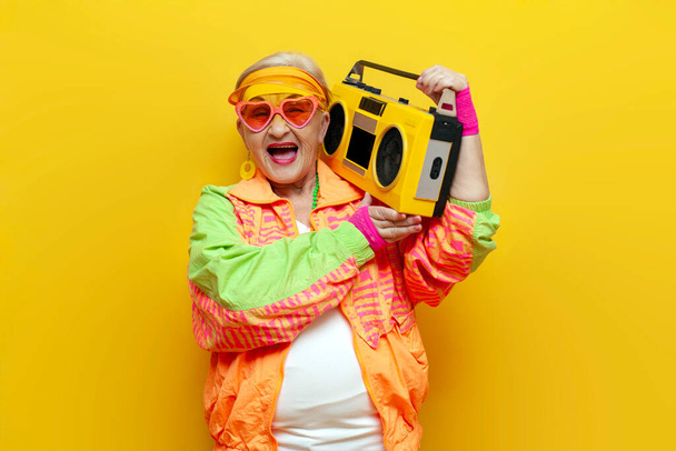 αστεία τρελή γιαγιά με hipster ρούχα ακούγοντας μουσική σε μαγνητόφωνο και τραγουδώντας σε κίτρινο απομονωμένο φόντο, ηλικιωμένη δροσερή γυναίκα χορεύει στο πάρτι και ουρλιάζει - Φωτογραφία, εικόνα