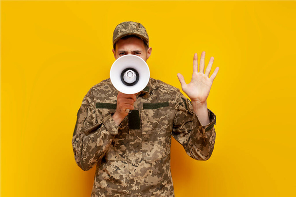 Ukrainian army soldier in pixel military camouflage uniform announces information into a megaphone and shouts on a yellow isolated background, Ukrainian military cadet speaks and warns into a - Photo, Image