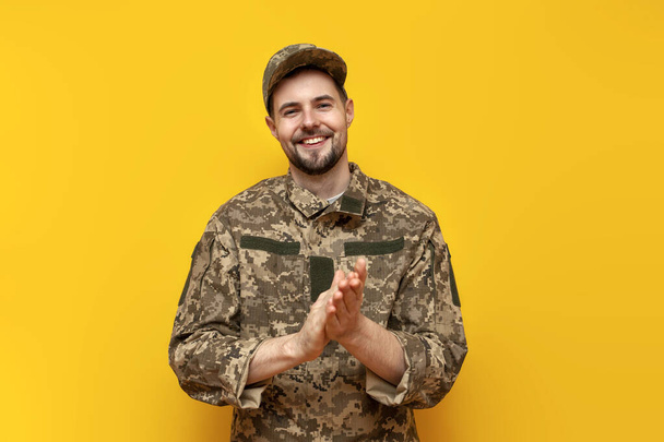 young Ukrainian army soldier in camouflage pixel uniform looks at the camera and applauds on a yellow isolated background, Ukrainian military cadet smiles and claps his hands - Photo, Image