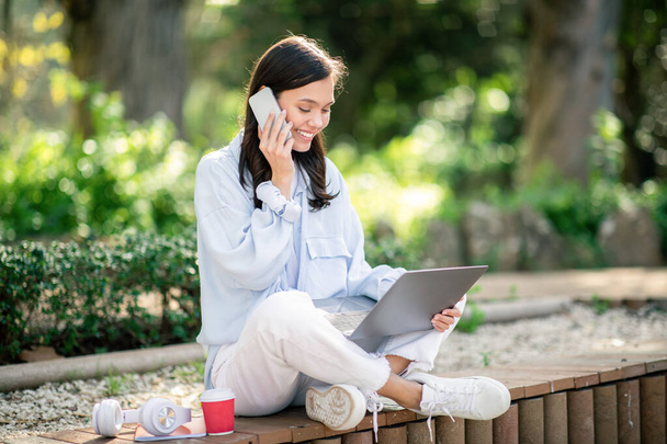Jeune femme européenne multitâche dans un cadre extérieur détendu, parlant joyeusement au téléphone tout en travaillant sur un ordinateur portable, avec une tasse de café et des écouteurs à proximité, à l'extérieur - Photo, image