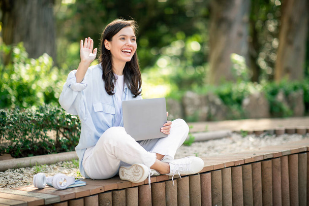 Joyful feliz jovem europeia acenando enquanto sentado em um banco de parque, segurando um laptop em seu colo, com um par de fones de ouvido e xícara de café no banco ao lado dela, fora - Foto, Imagem