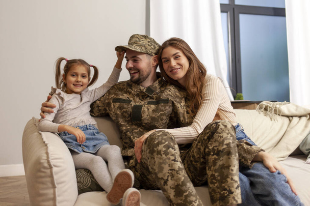 Ukraiński żołnierz armii w mundurze kamuflażu wrócił do domu do rodziny, kadeci wojskowi siedzi na kanapie z żoną i córką i uśmiecha się, dziecko przytula weteranów ojciec - Zdjęcie, obraz