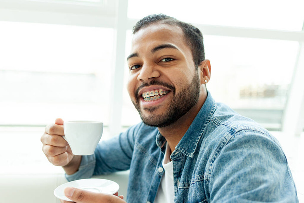 Afroamerikaner mit Hosenträgern trinkt Kaffee aus einer Tasse und lächelt in einem weißen Café, ein Mann mit Stoppeln trinkt Tee aus einem weißen Becher - Foto, Bild