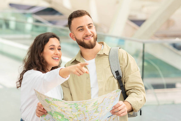 Hymyilevä pariskunta, jolla on kartta, näyttää navigoivan, naisen osoittaessa sijaintia, valoisassa lentokentän käytävässä. - Valokuva, kuva