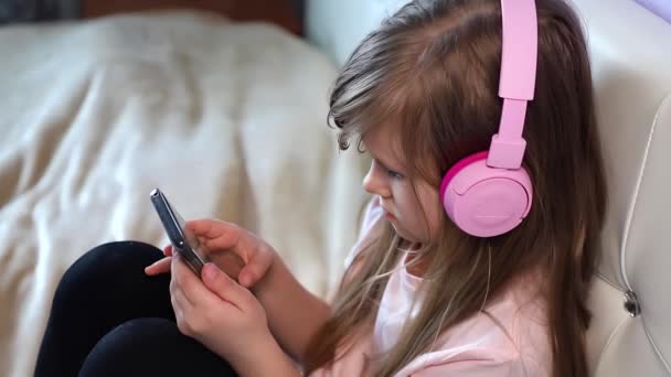Linda niña en los auriculares escuchar música y utilizar un teléfono inteligente sentado en una cama en casa, ángulo lateral de cámara lenta. Imágenes de alta calidad 4k - Metraje, vídeo